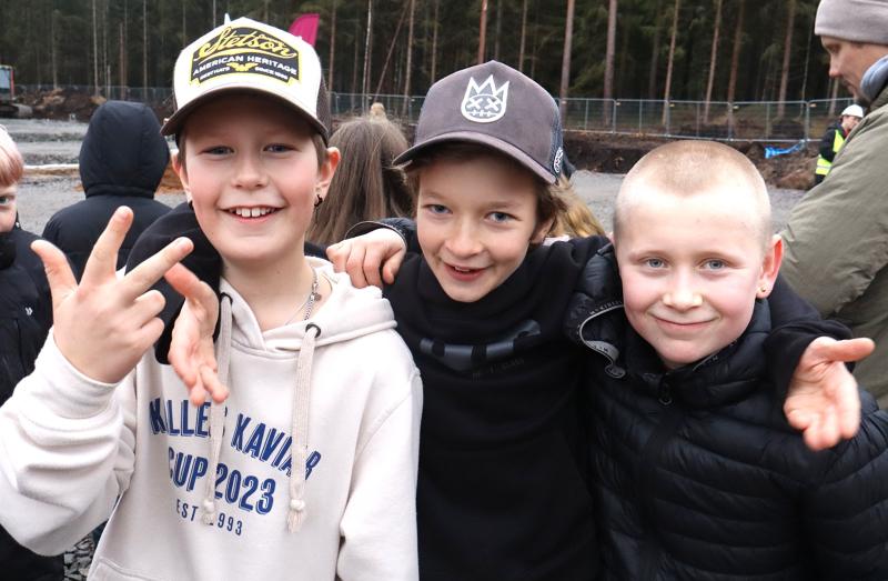 Tre pojkar i fritidsjackor och hattar står tätt tillsammans utomhus och ler mot kameran, med andra och träd i bakgrunden.