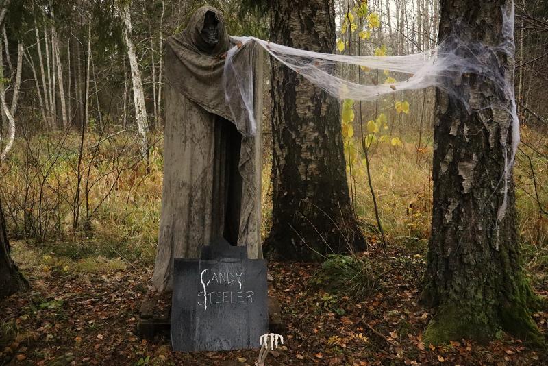 En Halloween-dekoration i form av en lieman med huva står i en skog och håller i falska spindelnät, med en skylt med texten 