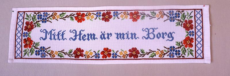 En rektangulär broderad skylt med färgglad blomsterbård och den svenska texten 