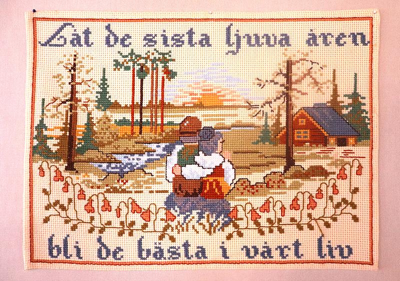 En korsstygnsbroschyr visar ett par som sitter på en bänk och tittar på solnedgången vid en sjö, med svensk text och ett hus, träd och blommor i bakgrunden.