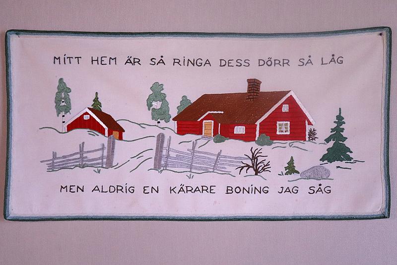 En dekorativ duk med en svensk fras och en illustration av två röda hus, ett staket, träd och en sten på en vit bakgrund.