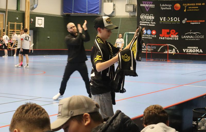 En pojke i sportkläder står i en gymnastiksal och håller i en svartgul halsduk, medan andra barn sitter i närheten och människor leker eller går i bakgrunden.