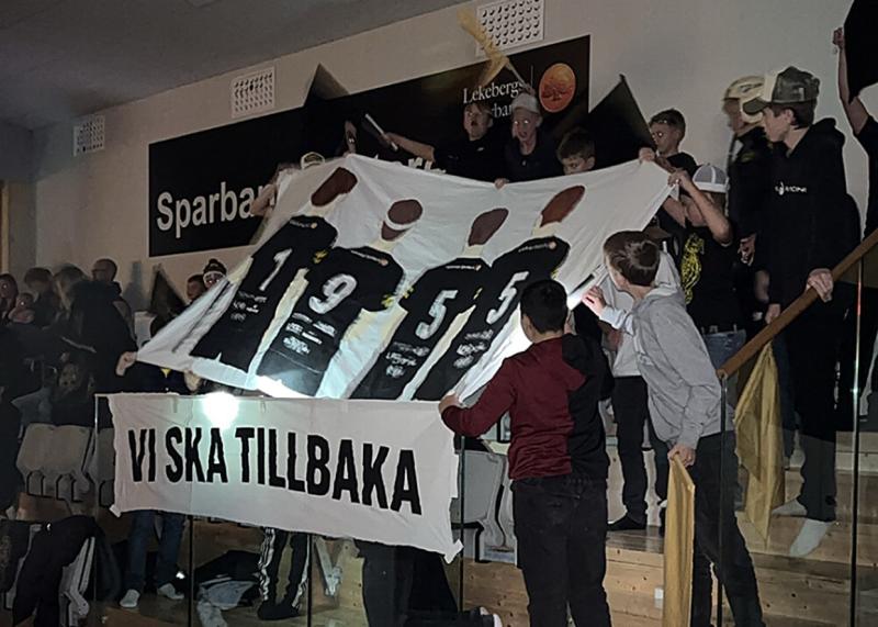 En grupp människor på en stadion står och håller i en stor banderoll med fyra fotbollsspelare och texten 