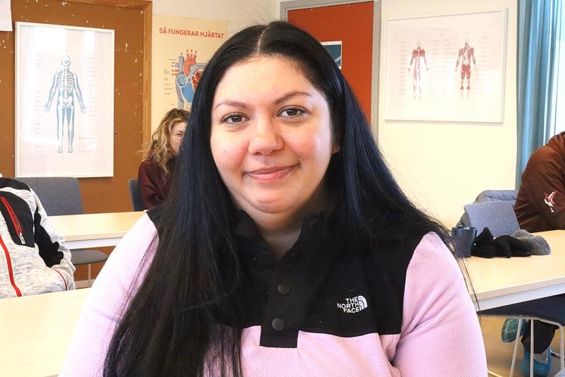 En kvinna med långt mörkt hår, klädd i en ljusrosa och svart North Face-jacka, sitter i ett klassrum med anatomiska affischer på väggarna och andra människor i bakgrunden.