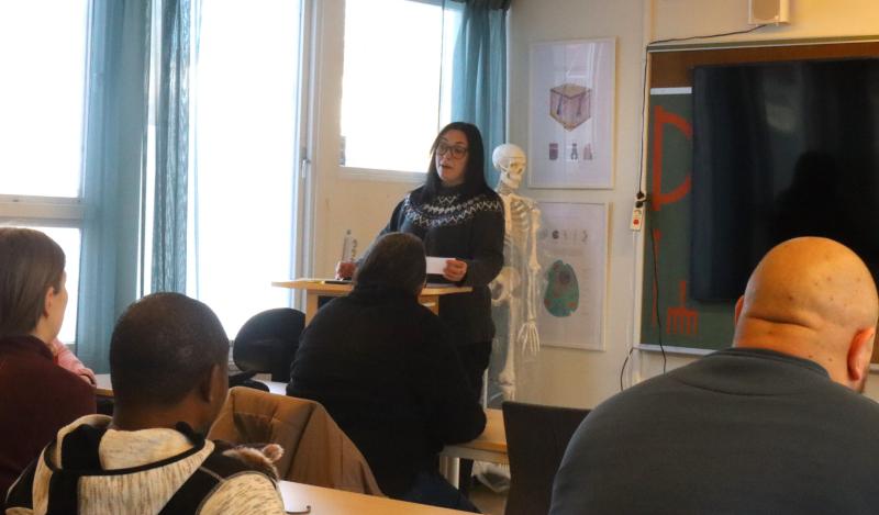 En kvinna står vid ett podium och talar till en grupp sittande personer i ett klassrum, med utbildningsaffischer och en skelettmodell i bakgrunden.