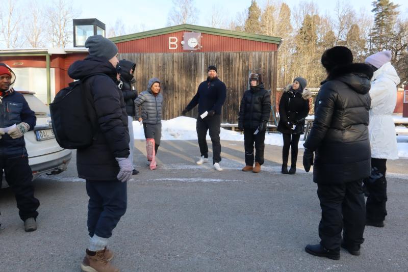 En grupp människor klädda i vinterkläder står i en cirkel utomhus på en asfalterad yta nära en röd byggnad en kall, solig dag.
