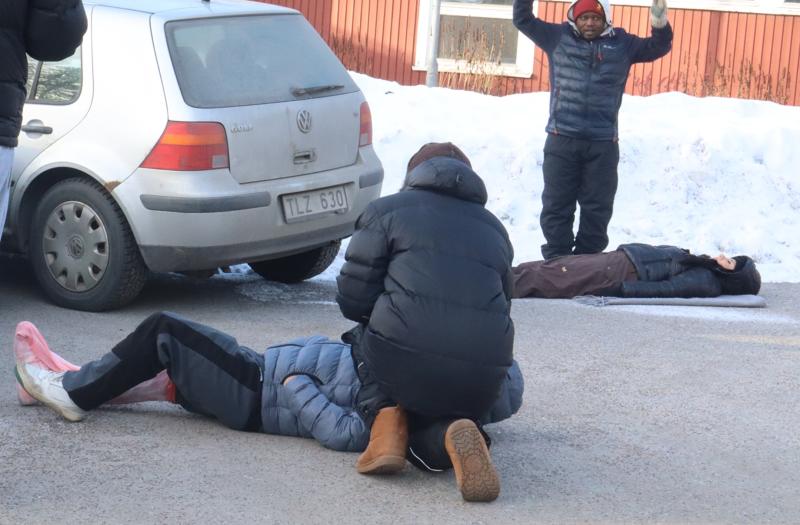 Två personer ligger på den snöiga marken nära en bil, den ena får hjälp av en annan person; en fjärde person står i närheten med händerna uppsträckta.