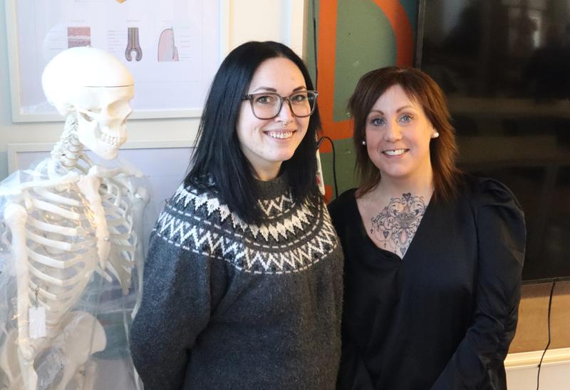 Två kvinnor står inomhus bredvid en modell av ett mänskligt skelett och ler mot kameran. Den ena bär glasögon och en grå tröja, den andra en svart topp med en synlig tatuering på bröstet.