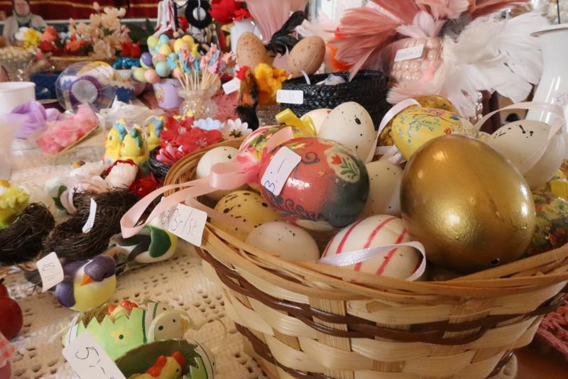 En flätad korg fylld med dekorerade påskägg ligger på ett bord bland olika färgglada hantverk och prydnadsföremål, alla märkta med en prislapp.