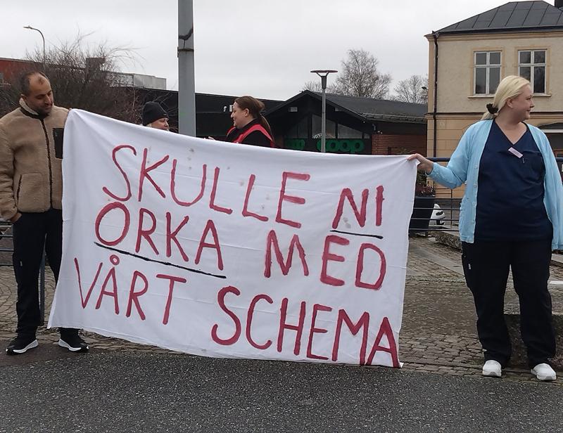 Fyra personer står utanför och håller en stor handskriven banderoll med texten 