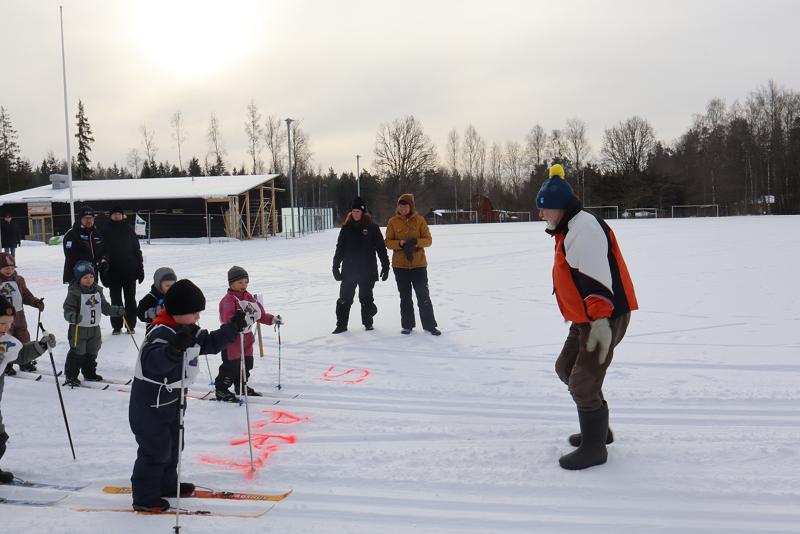 En grupp barn på skidor samlas vid en snöig startlinje med vuxna som övervakar dem; en vuxen i orange jacka står vänd mot barnen.