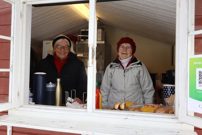 Två äldre kvinnor står bakom ett servicefönster och ler. På disken finns kryddor, termosar och förpackade matvaror. Miljön ser ut att vara ett litet kök eller ett matstånd.