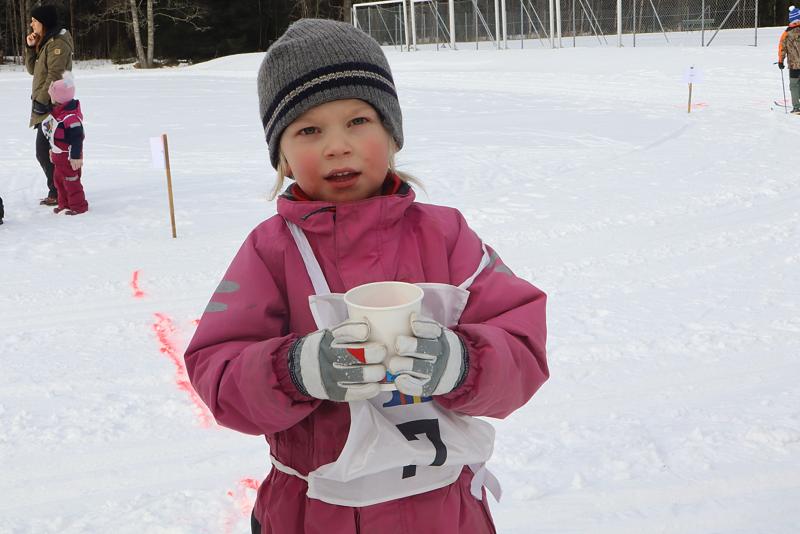 Ett barn i rosa snödräkt och grå mössa håller i en pappersmugg när det står på ett snöigt fält; andra människor syns i bakgrunden.