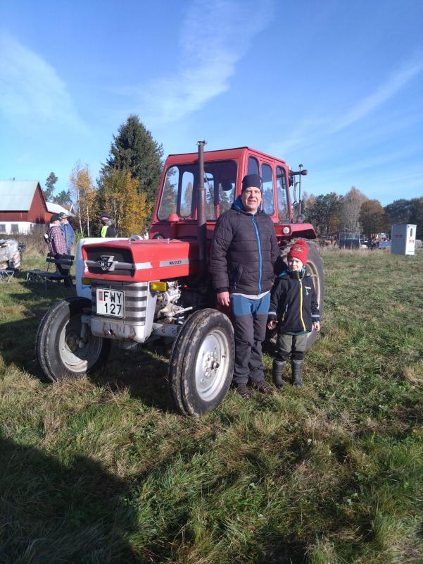 Två personer står bredvid en röd Massey Ferguson-traktor på ett gräsbevuxet fält, med träd och byggnader i bakgrunden.